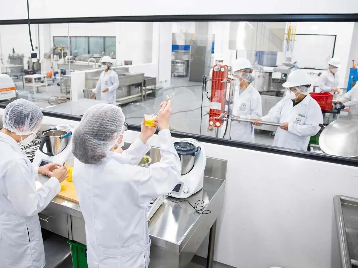 Operators in hairnets and lab coats running a stainless food-processing line behind a viewing window
